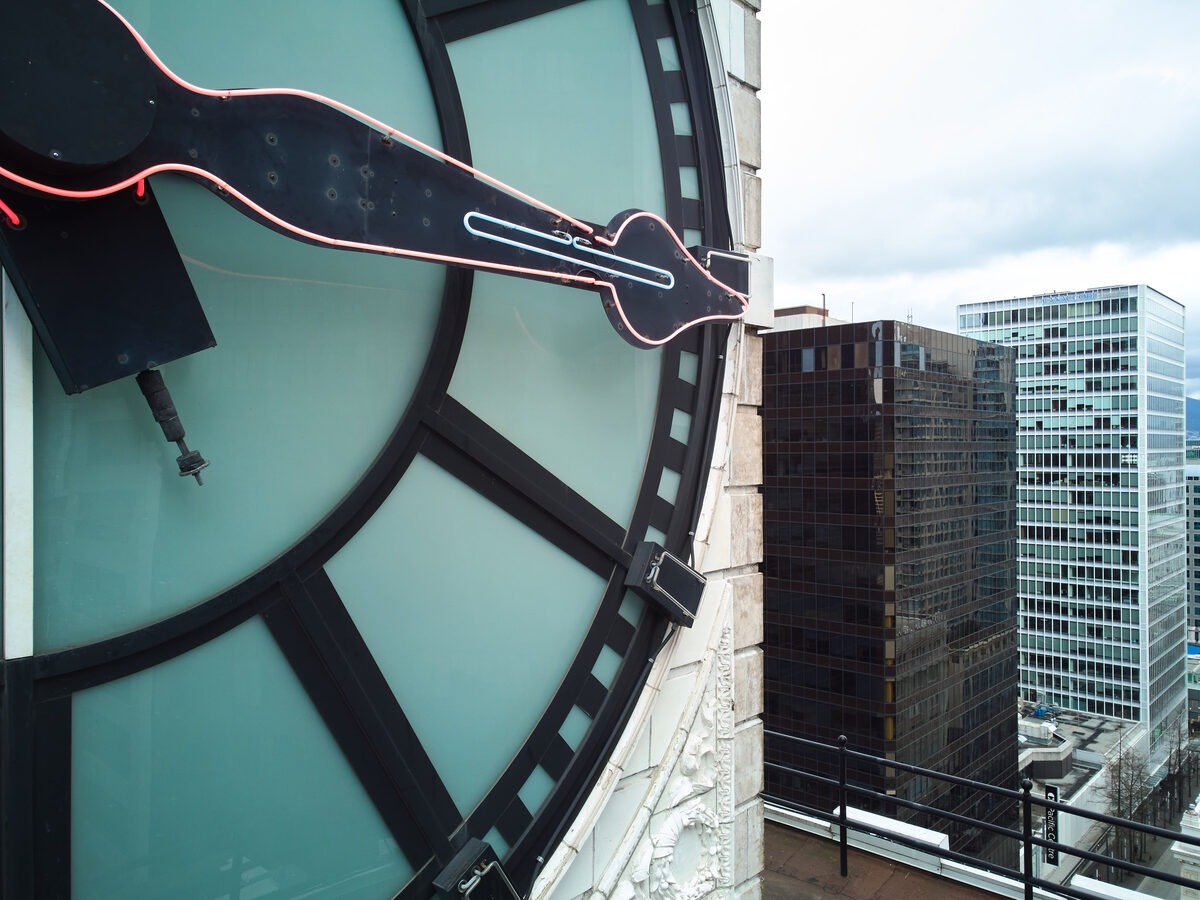 Neon-outlined clock hands glowing against the translucent glass face with the city beyond
